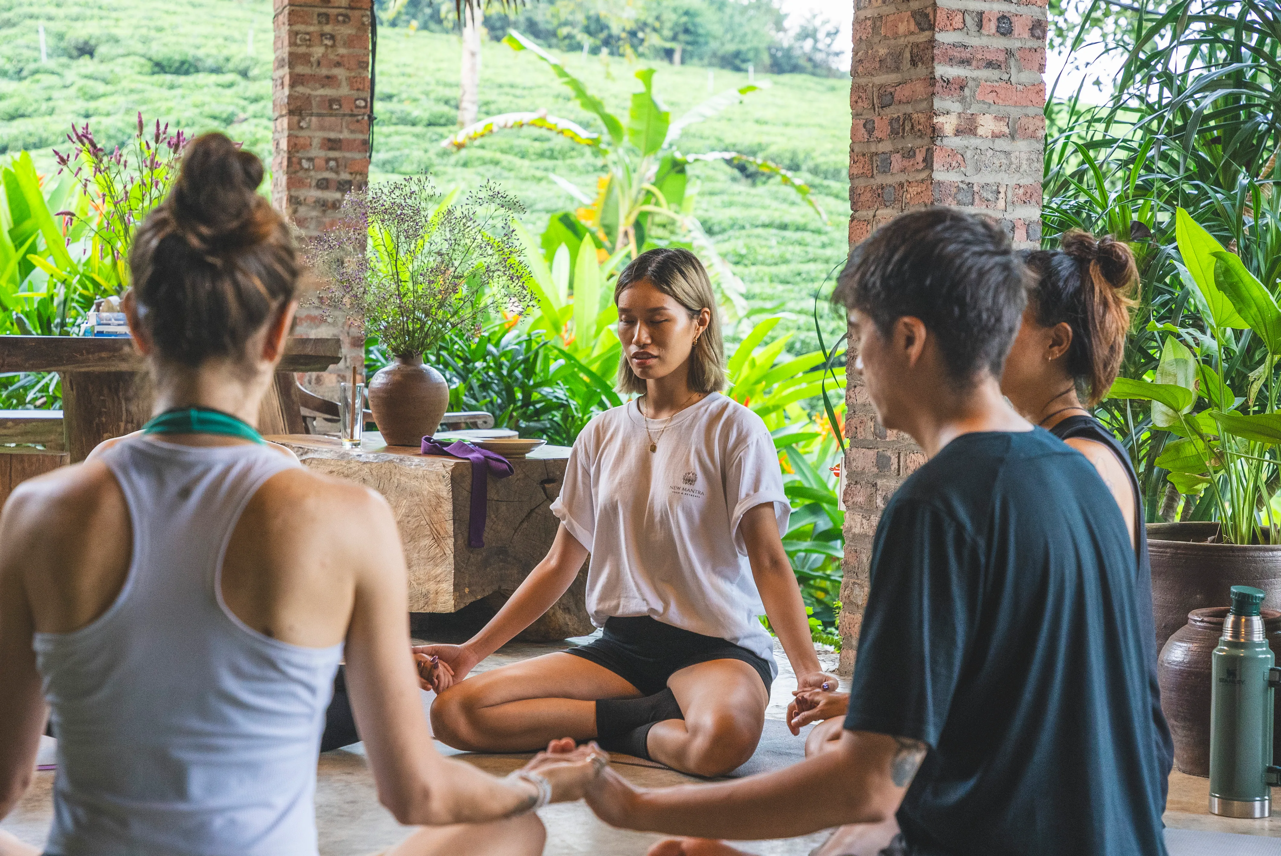 A small group sitting together in a circle during a retreat, hands joined at the centre, surrounded by lush greenery